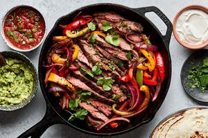 steak fajitas and peppers in a cast iron skillet sprinkled with cilantro surrounded by salsa, guacamole and corn tortillas