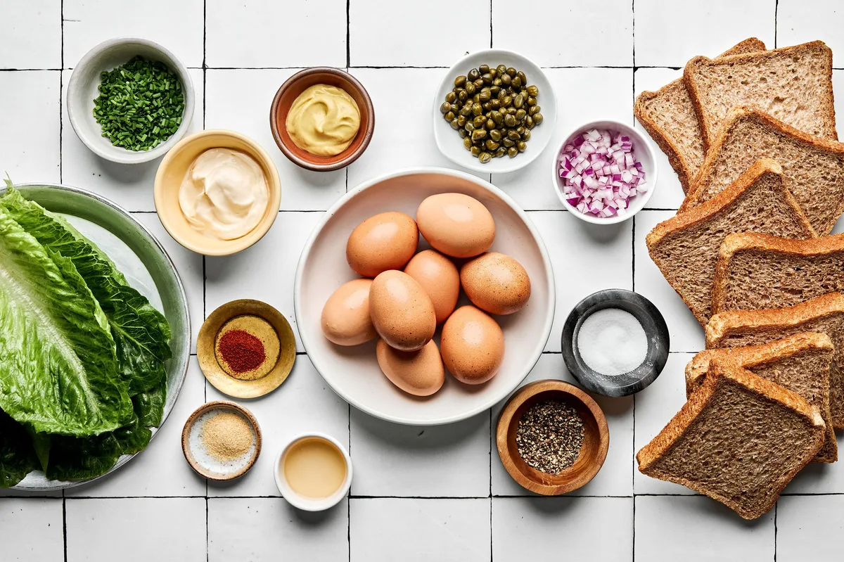 ingredients for egg salad sandwiches laid out: bread, boiled eggs, dijon, mayo, pickle juice, chives, spices and romaine