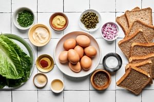 ingredients for egg salad sandwiches laid out: bread, boiled eggs, dijon, mayo, pickle juice, chives, spices and romaine