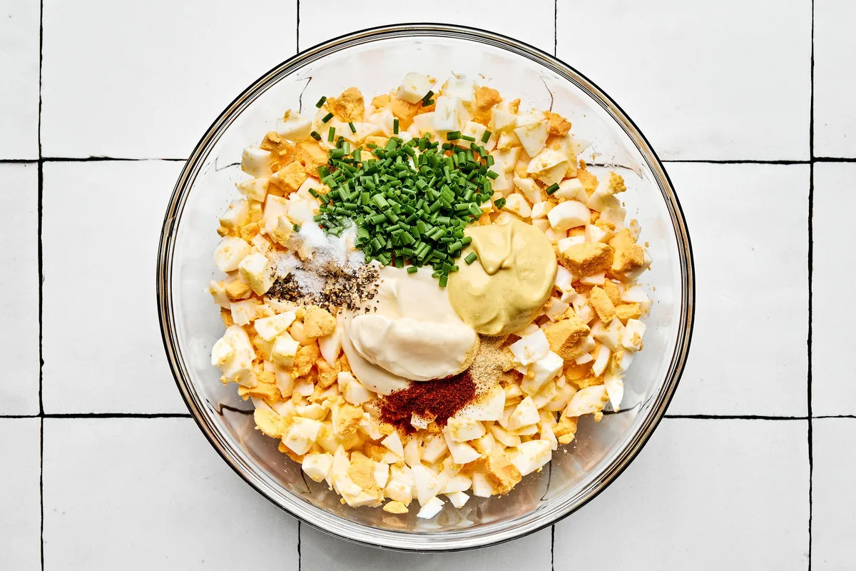 ingredients for egg salad being mixed in a bowl including chopped boiled egg, mayo, dijon, pickle juice chives and spices