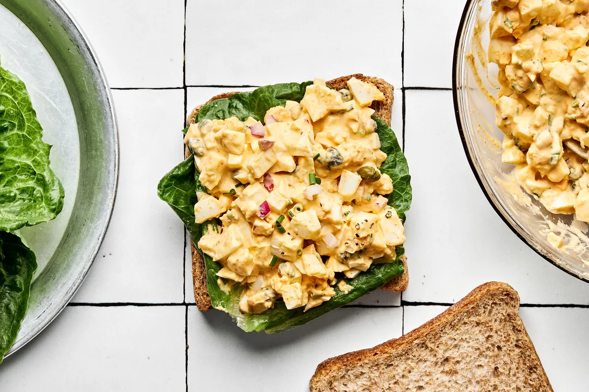 the best egg salad sandwich being assembled on soft bread topped with romaine lettuce and creamy egg salad filling