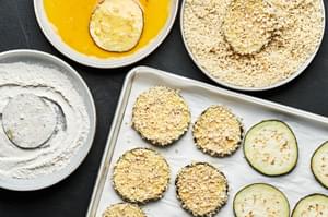 Eggplant slices dipped in egg, flour and panko on a parchment lined baking sheet surrounded by three bowls of ingredients.
