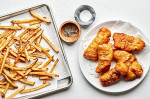 homemade cod fish and chips on paper lined plates next to bowls of salt and pepper for seasoning