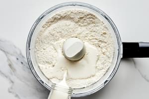 heavy cream being poured into a food processor with flour, baking powder, sugar, and salt for fresh berry cobbler