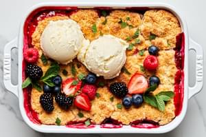 fresh berry cobbler in a 9x13 baking dish topped with 2 scoops of vanilla ice cream, additional fresh berries and fresh mint