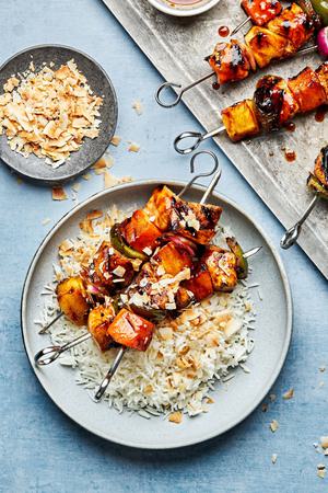 hawaiian chicken kabobs on top of coconut rice topped with flaked coconut next to a baking sheet of Hawaiian chicken kabobs