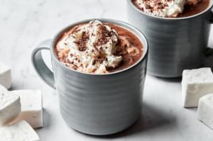 2 mugs of homemade hot chocolate topped with whipped cream and chocolate chips served with marshmallows