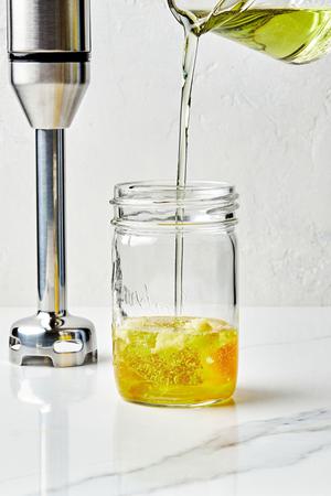 an egg, Garlic, Dijon, salt, Lemon juice and zest in a mason jar with grapeseed oil being poured in to make homemade mayo