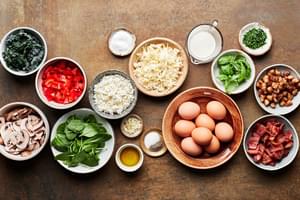 eggs, milk, shredded cheese, chopped cooked bacon, chopped vegetables, spices and basil in bowls to make a homemade frittata