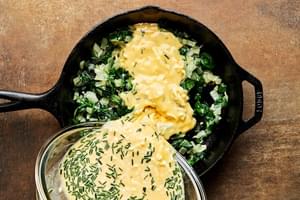 egg, milk, cheese and seasoning mixture being poured into a cast iron skillet with cooked veggies for making a frittata