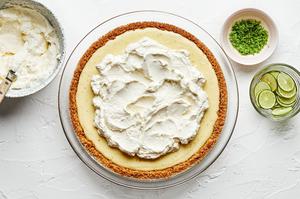 a homemade key lime pie topped with whipped cream next to bowls of extra whipped cream, lime zest and sliced limes