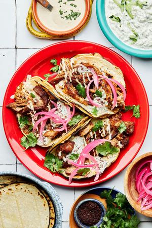 three lamb tacos on a plate topped with cilantro, tahini, tzatziki and pickled onions surrounded by plates of garnishes