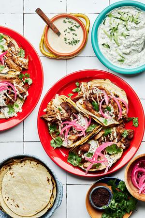 three lamb tacos on a plate topped with cilantro, tahini, tzatziki and pickled onions surrounded by plates of garnishes