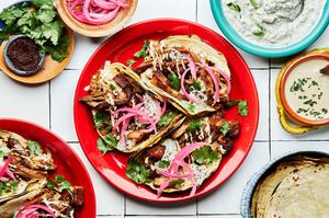 three lamb tacos on a plate topped with cilantro, tahini, tzatziki and pickled onions surrounded by plates of garnishes