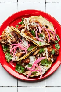three homemade lamb tacos on a plate topped with cilantro, tahini and tzatziki sauces, sumac and pickled onions