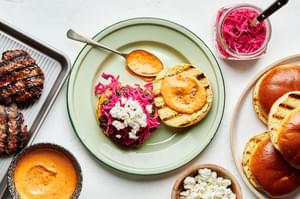 an open lamb burger topped with picked red cabbage and chèvre with Harrisa mayo being spread next to bowls of more fixings