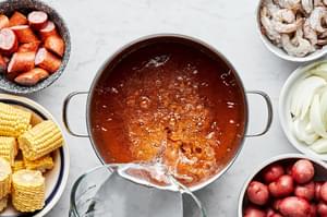 water being poured into a large pot with chicken stock, smashed garlic and old bay seasoning to make a low country boil