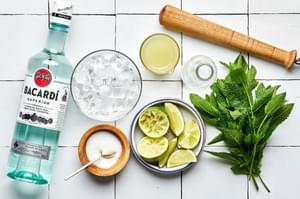 a bottle of rum, glass of ice, lime juice, sugar, mint and lime wedges in bowls and a muddler to make a mojito
