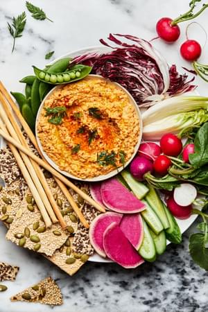 homemade nutty carrot dip in a bowl on top of a platter surrounded with raw peas, cucumbers, radishes and crackers