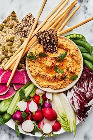 homemade nutty carrot dip in a bowl on top of a platter surrounded with raw peas, cucumbers, radishes and crackers