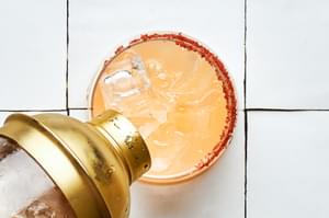 A Paloma being poured from a cocktail shaker into a salt lined glass full of ice made with tequila, grapefruit & lime juices
