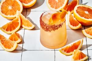 paloma in a salt rimmed glass full of ice garnished with a grapefruit wedge surrounded by wedges of grapefruit on the counter