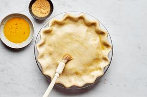 an unbaked peach pie being brushed with egg wash on the top crust