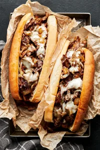 Two homemade Philly cheesesteak sandwiches on hoagie rolls wrapped in parchment paper on a baking sheet