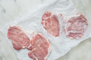 boneless pork chops on parchment paper with plastic wrap on top ready to be pounded with a mallet for pork schnitzel