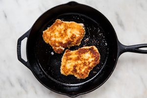 2 pork chops dredged in flour, egg and plain breadcrumbs being fried in vegetable oil in a frying pan for pork schnitzel