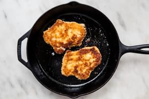 2 pork chops dredged in flour, egg and plain breadcrumbs being fried in vegetable oil in a frying pan for pork schnitzel