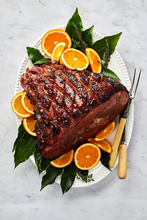 a homemade roasted garlic honey baked ham on a serving platter surrounded by bay leaves and fresh orange slices