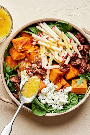 A Roasted Sweet Potato Salad topped with candied pecans, goat cheese and apples being drizzled with Orange Vinaigrette