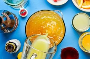 Glass pitcher pouring pineapple juice into a large pitcher of rum punch surrounded by bitters, lime juice, pineapple juice.
