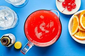 rum punch in a glass pitcher, surrounded by ice, oranges, bitters and Maraschino cherries in small bowls.