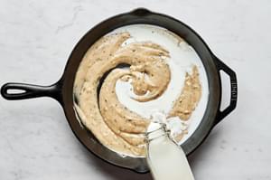 milk being poured into a skillet with butter, flour and garlic that have been cooked and thickened