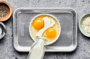 a butter greased ramekin on top of a baking sheet with 2 cracked eggs in it and cream being poured in