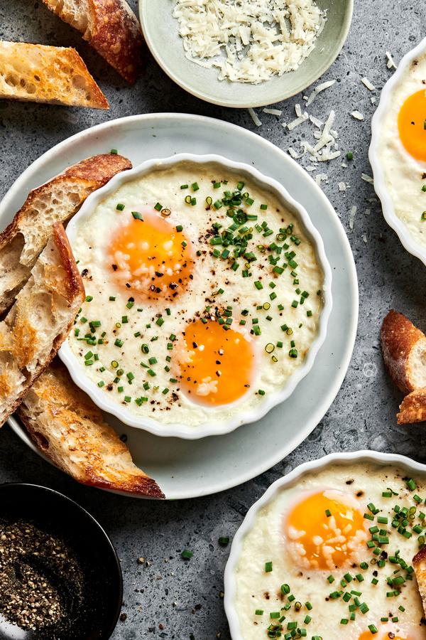 ramekins of shirred eggs garnished with chopped chives and served with crusty bread next to a bowl of parmesan