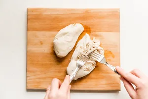 2 cooked chicken breasts on a cutting board being shredded with two forks that were boiled in water with salt