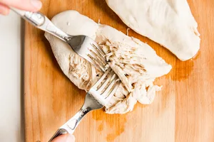 two cooked chicken breasts on a cutting board being shredded with 2 forks