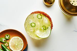 a spicy margarita in a rocks glass with a tajín seasoning rim next to a plate of lime wedges and serrano peppers for garnish