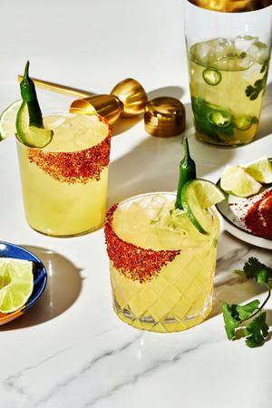 2 spicy margaritas in rocks glasses next to a cocktail shaker and a plate of tajín seasoning and limes garnishes