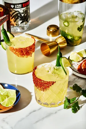 2 spicy margaritas in rocks glasses next to a cocktail shaker and a plate of tajín seasoning and limes garnishes
