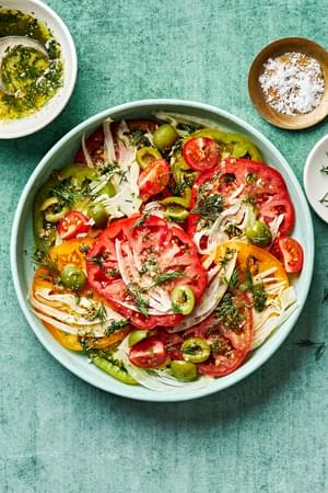 tomato salad on a platter topped with flaky salt and fresh dill surrounded by bowls of extra dressing, flaky salt and dill