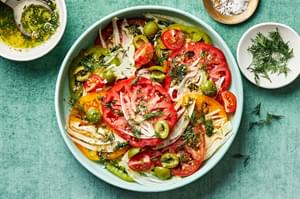 tomato salad on a platter topped with flaky salt and fresh dill surrounded by bowls of extra dressing, flaky salt and dill