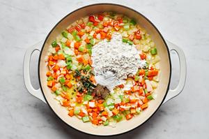flour and spices being added to a skillet with onion, carrots, celery and garlic that were cooked in melted butter