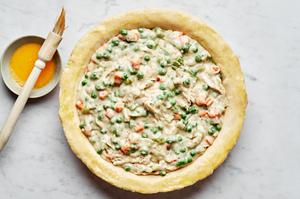 pie crust filled with turkey pot pie filling next to a bowl of egg wash and a pastry brush for brushing the pie crust