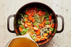 chicken broth being poured into a pot with carrots, onion, celery, garlic and butter for turkey soup