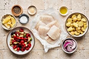white fish, potatoes, spices, red onion and a bowl of garlic, marinated tomatoes & olives laid out to make baked fish