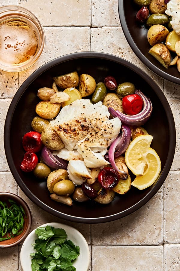 Fish baked on a sheet pan with potatoes, olives, onion, garlic & tomatoes beautifully presented in a bowl with lemon slice